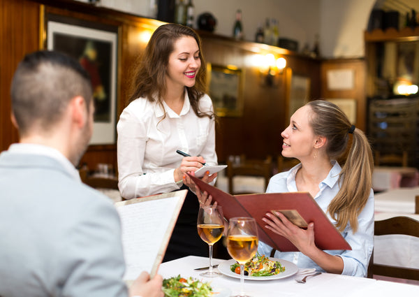 Eine Kellnerin nimmt mit einem Lächeln auf den Lippen die Bestellung von einem Parr auf. Auf dem Tisch stehen zwei Weingläser und zwei Salate.
